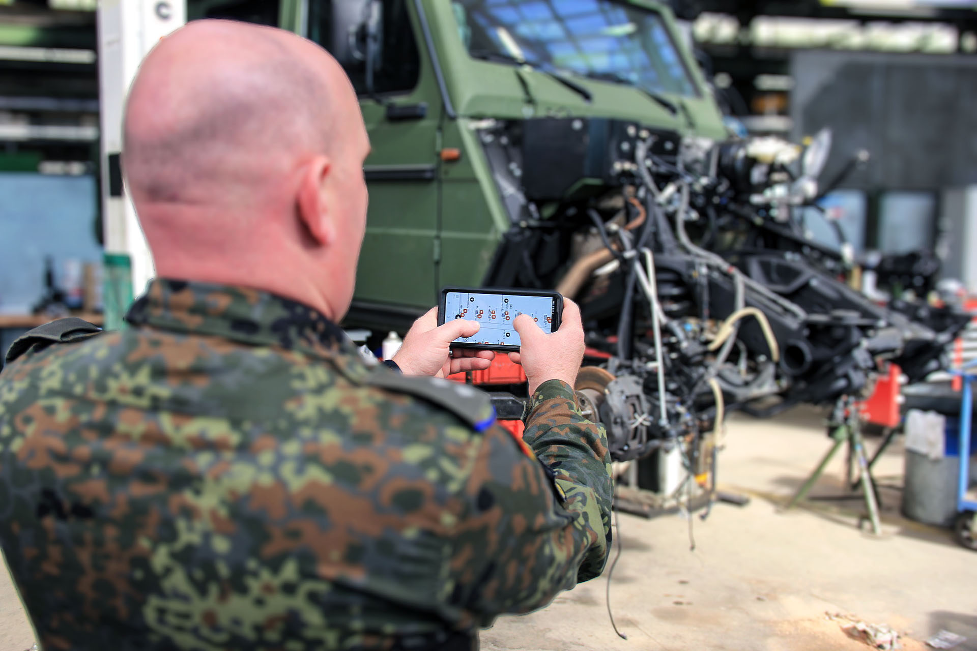 Soldat nutzt Smartphone vor geöffnetem Militärfahrzeug in Werkhalle