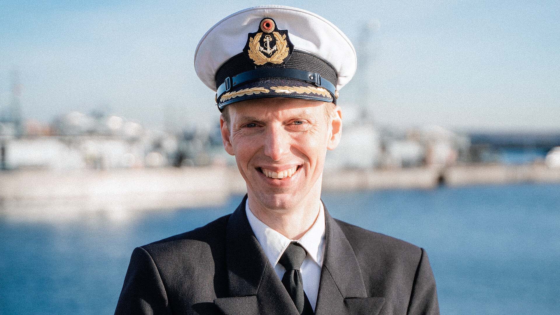 Korvettenkapitän Patrick O’Keeffe steht in Marineuniform vor Hafen mit Schiff im Hintergrund