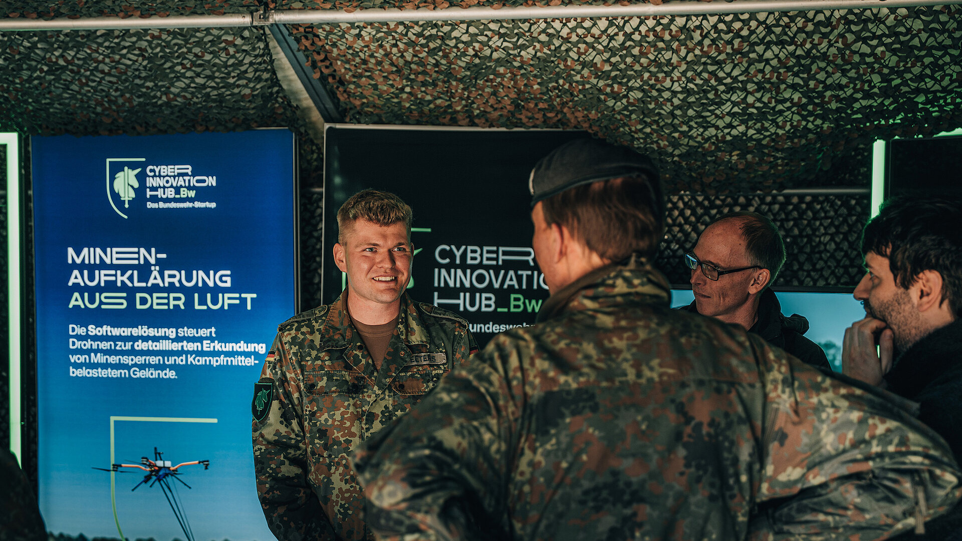 Mehrere Personen sprechen an einem Informationsstand, im Hintergrund Displays mit der Aufschrift ‚Cyber Innovation Hub Bw‘ und Textinhalten