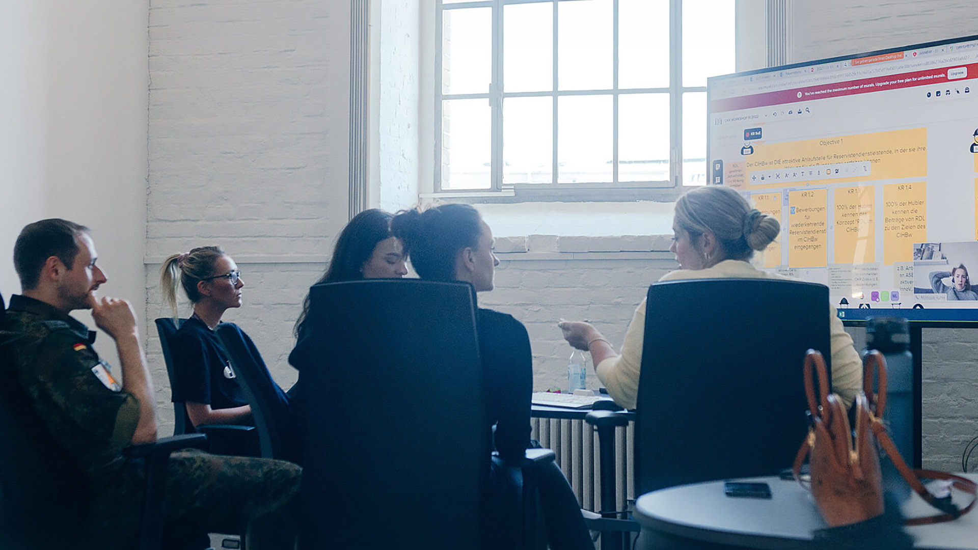 Mehrere Personen sitzen in einem Innenraum am Tisch vor Bildschirm