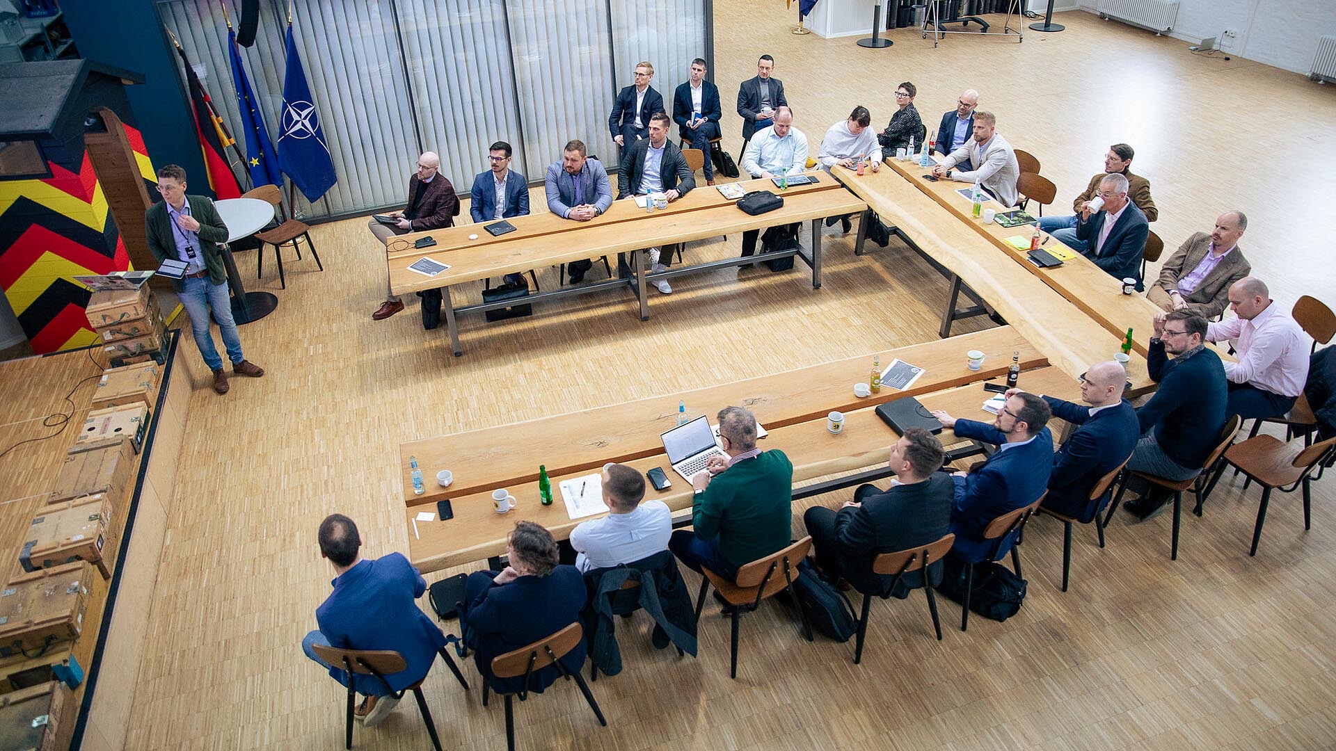 Teilnehmende sitzen an langen Tischen und verfolgen Präsentation im Raum