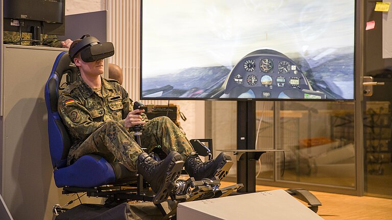 Ein Soldat sitzt im VR-Segelflugsimulator und fliegt. Dahinter ein Bildschirm.