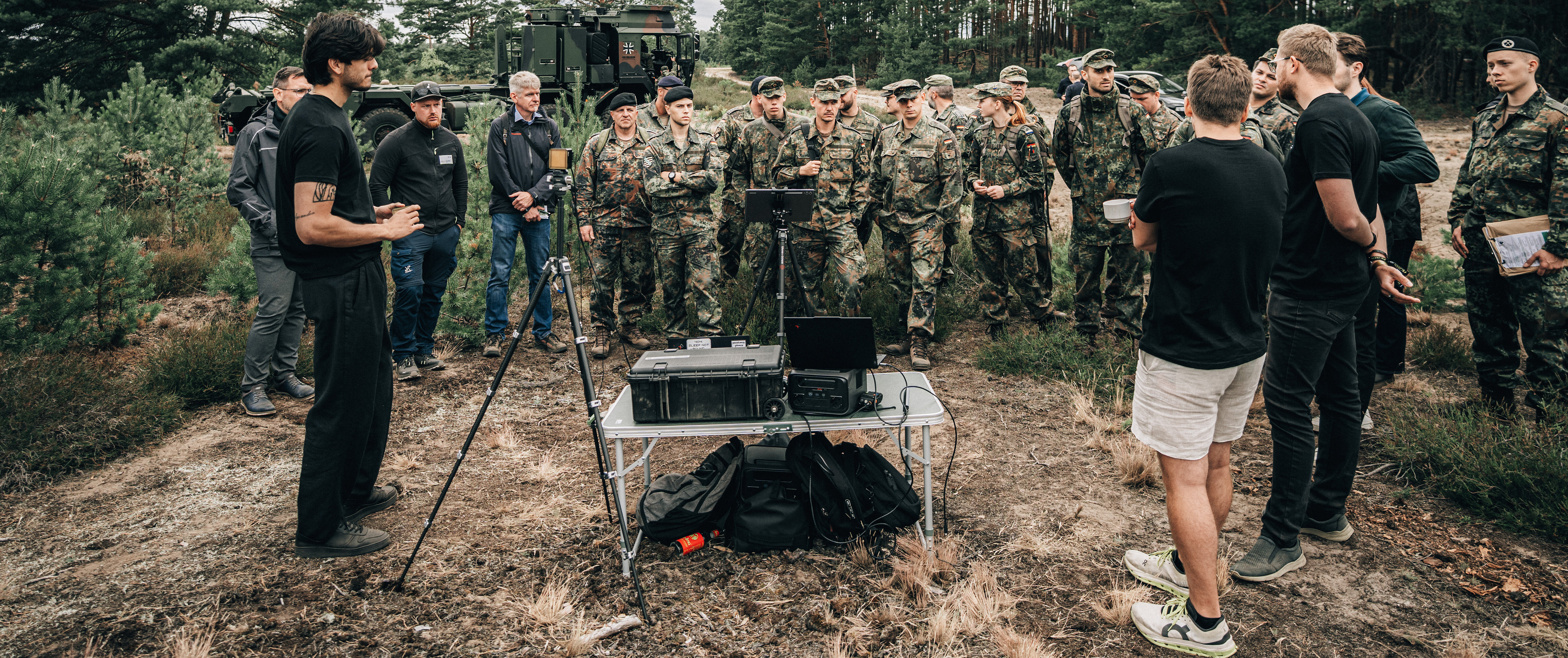 Eine Gruppe Soldatinnen und Soldaten steht auf einer Lichtung und lässt sich von einem Zivilisten einen Prototypen vorführen.