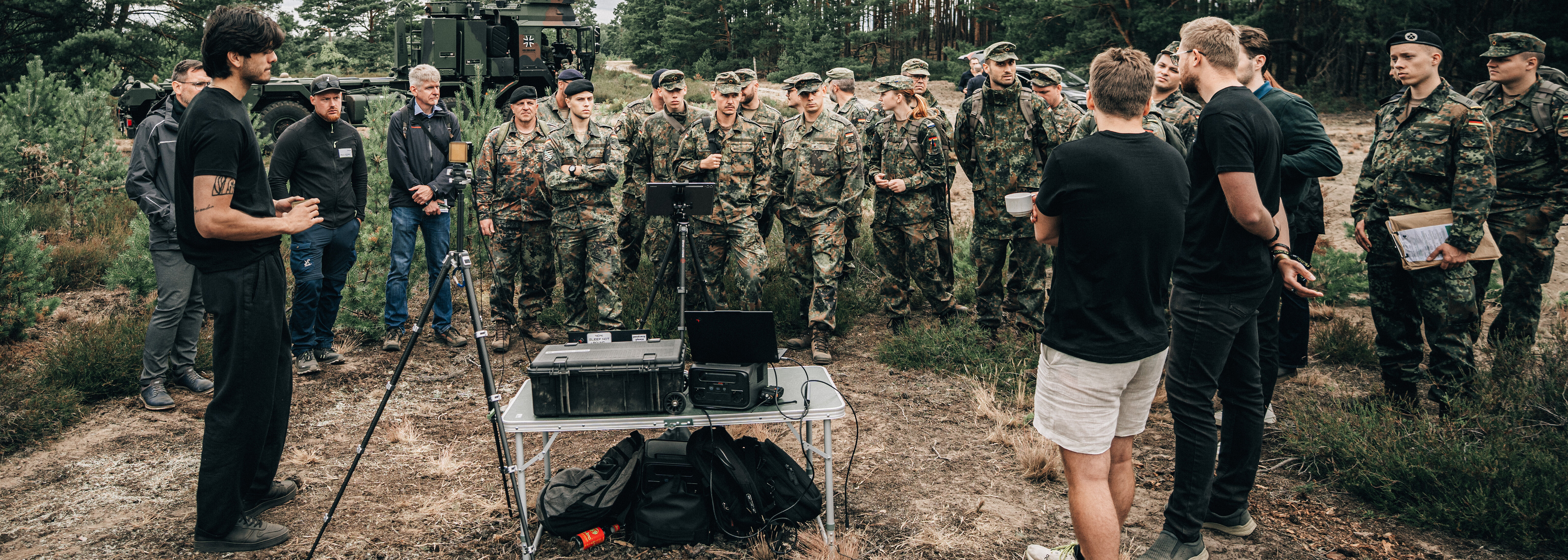 Eine Gruppe Soldaten steht auf einer Lichtung im brandenburgischen Kiefernwald. Im Vordergrund präsentieren Mitarbeitende eines Startup ihr Produkt.
