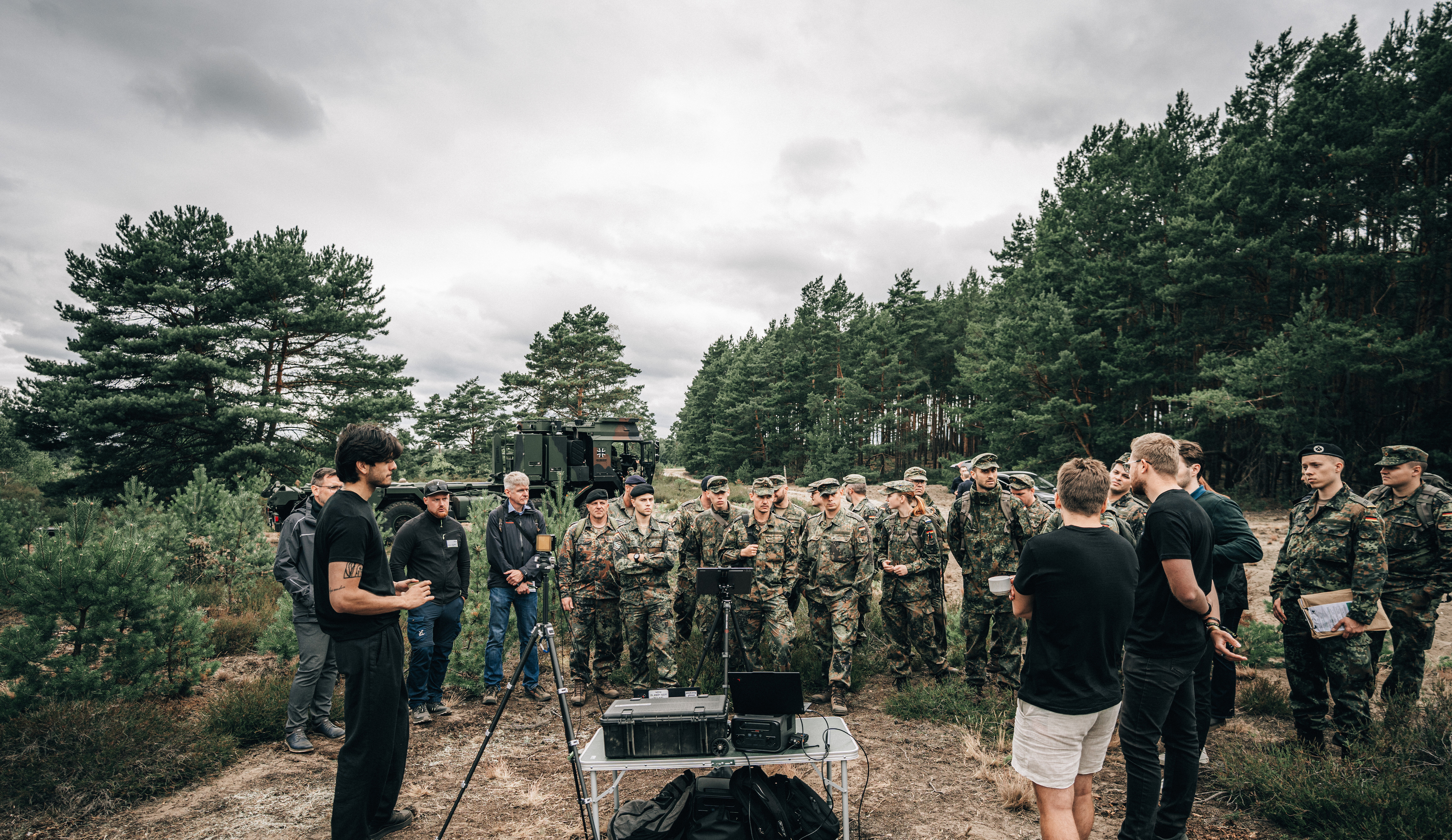 Eine Gruppe Soldaten steht auf einer Lichtung im brandenburgischen Kiefernwald. Im Vordergrund präsentieren Mitarbeitende eines Startup ihr Produkt.