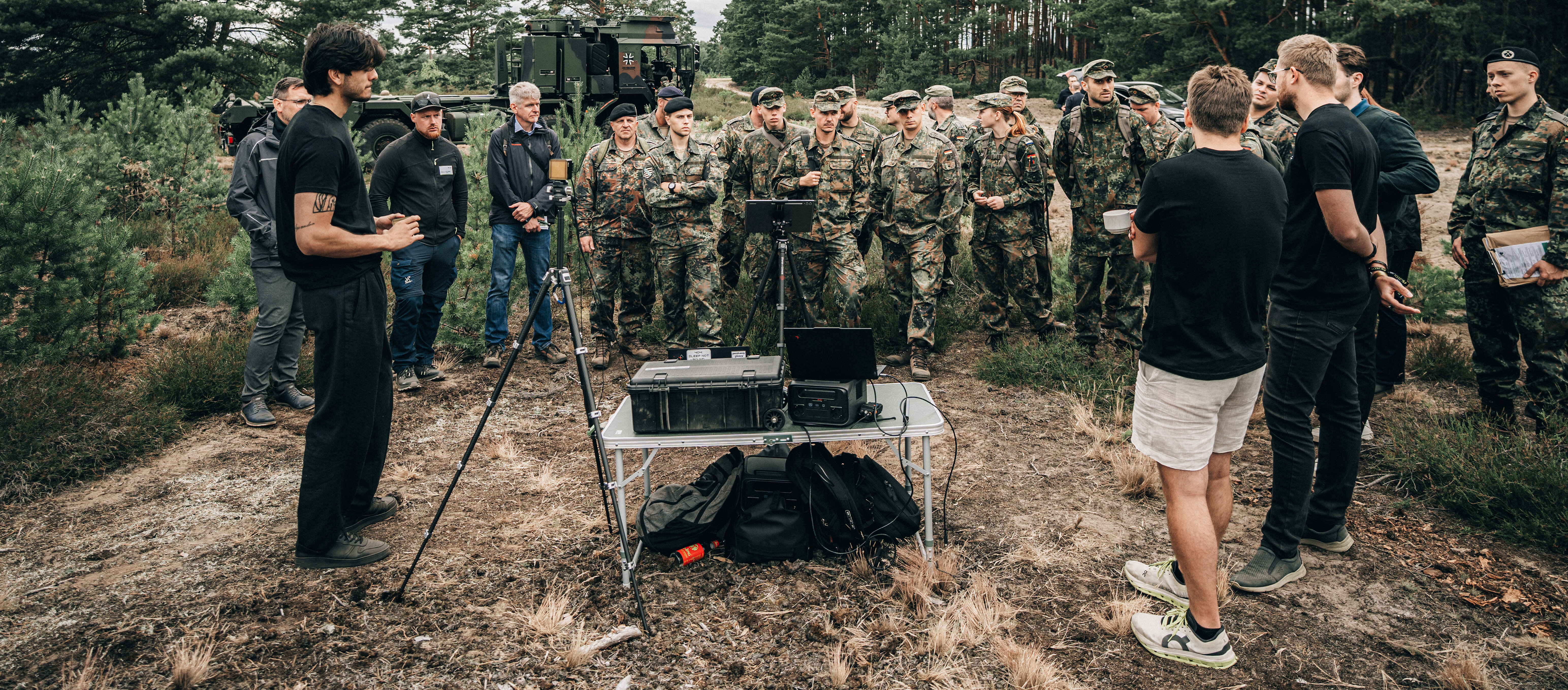Eine Gruppe Soldatinnen und Soldaten steht auf einer Lichtung und lässt sich von einem Zivilisten einen Prototypen vorführen.