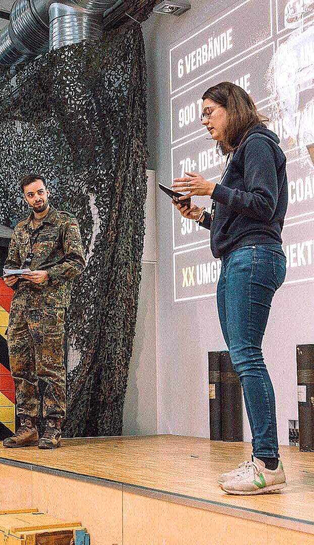 Zwei Personen präsentieren Projekt vor Bühne mit Flaggen im Hintergrund