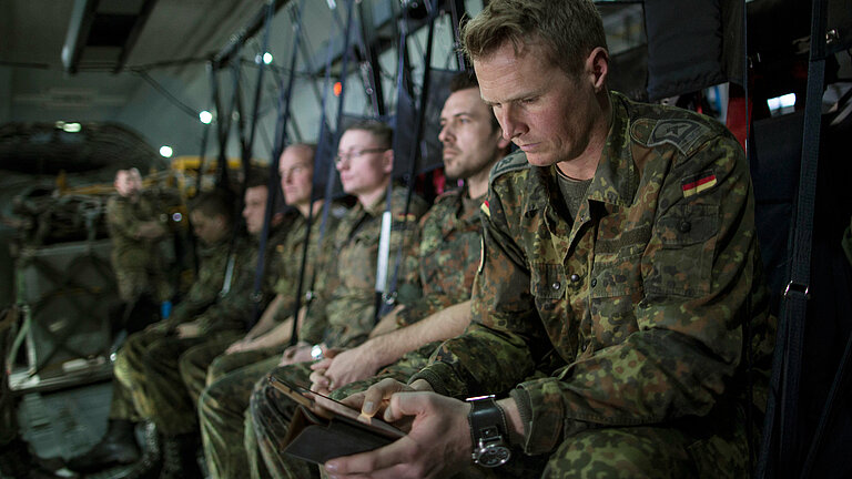 Mehrere Soldaten sitzen in einem Flugzeug, der vorderste hat ein Smartphone in der Hand.