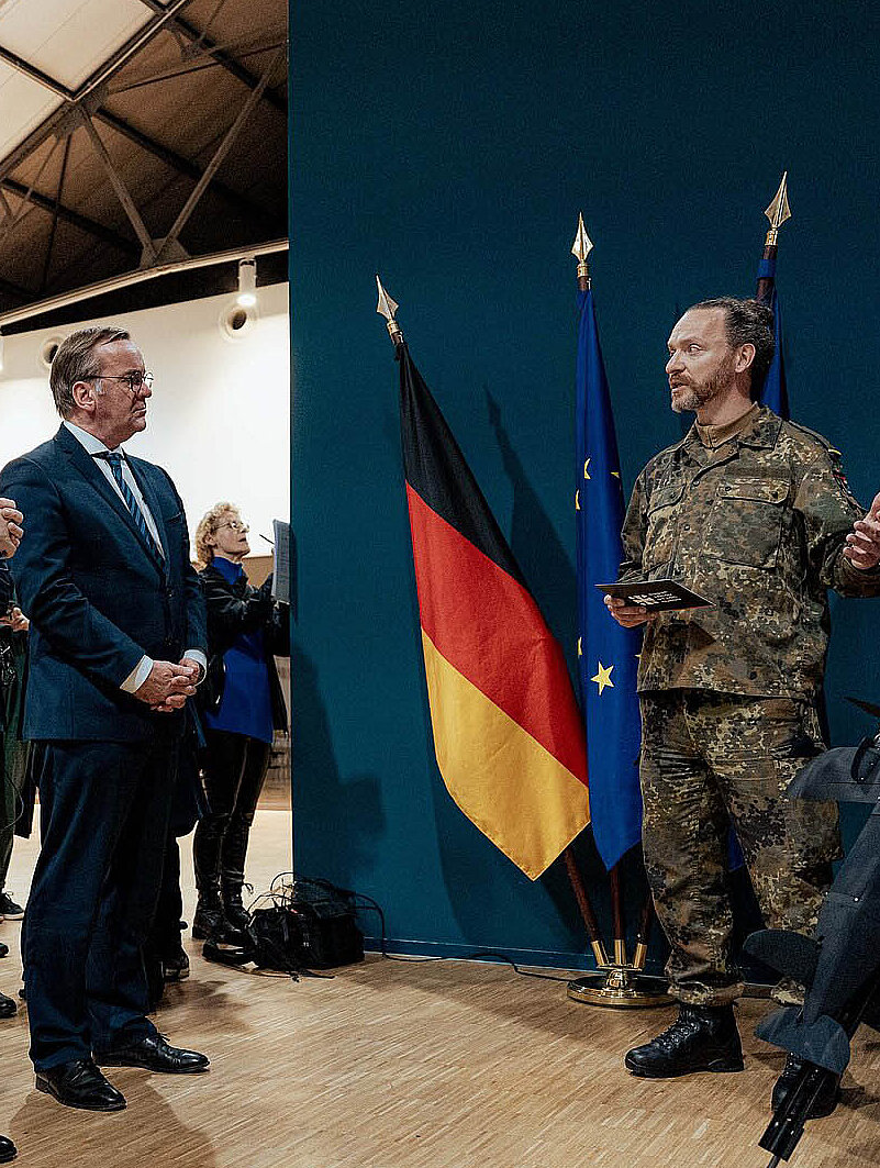 Soldat präsentiert Drohne vor Gruppe und Flaggen im Innenraum