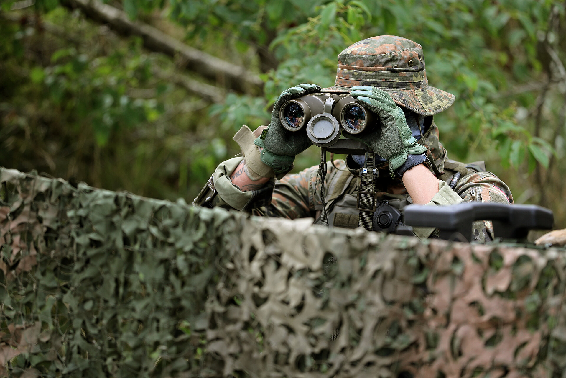 Ein Soldat blickt mit einem Fernglas über eine Absperrung. 