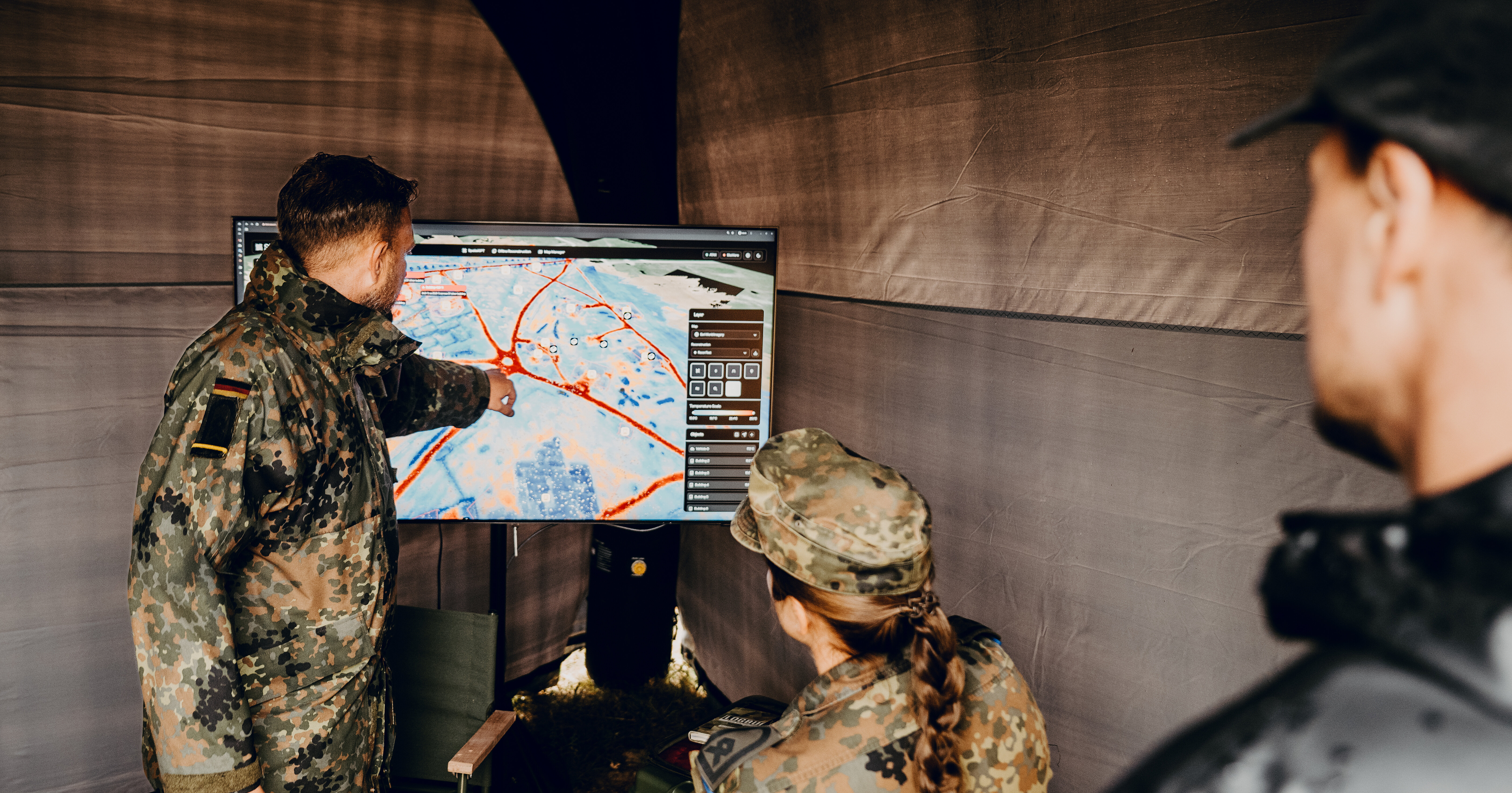 Zwei Soldaten und ein Zivilist stehen in einem Zelt vor einem Bildschirm, auf dem eine virtuelle Darstellung des Geländes zu sehen ist.