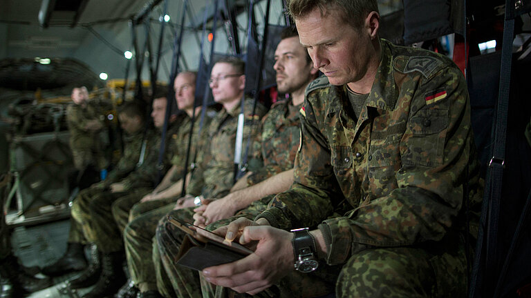 Several soldiers are sitting in a plane, the one in front has a smartphone in his hand.