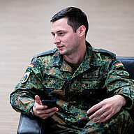 Christian Wendland sitzt in Uniform in Sessel und hält Smartphone im Gespräch