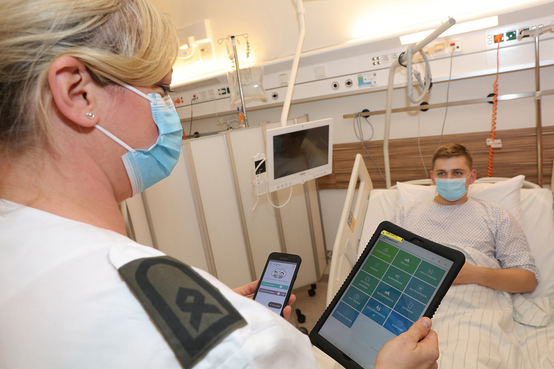 Eine Pflegerin geht mit Smartphone und Tablet in der Hand zu einem Patienten.
