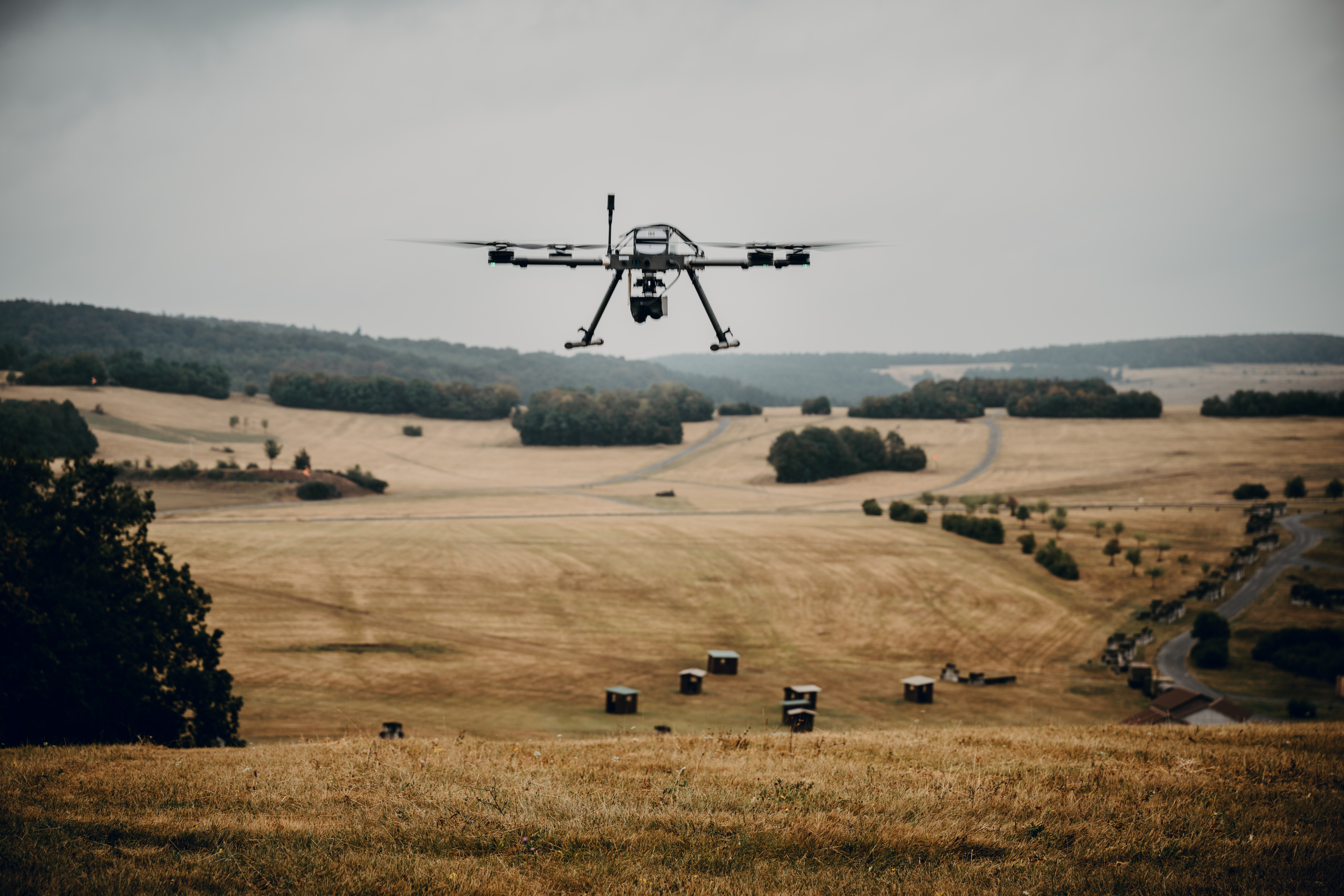Eine Drohne fliegt an einem regnerischen Tag über eine große Freifläche mit verdorrtem Gras und einem Üb-Dorf.