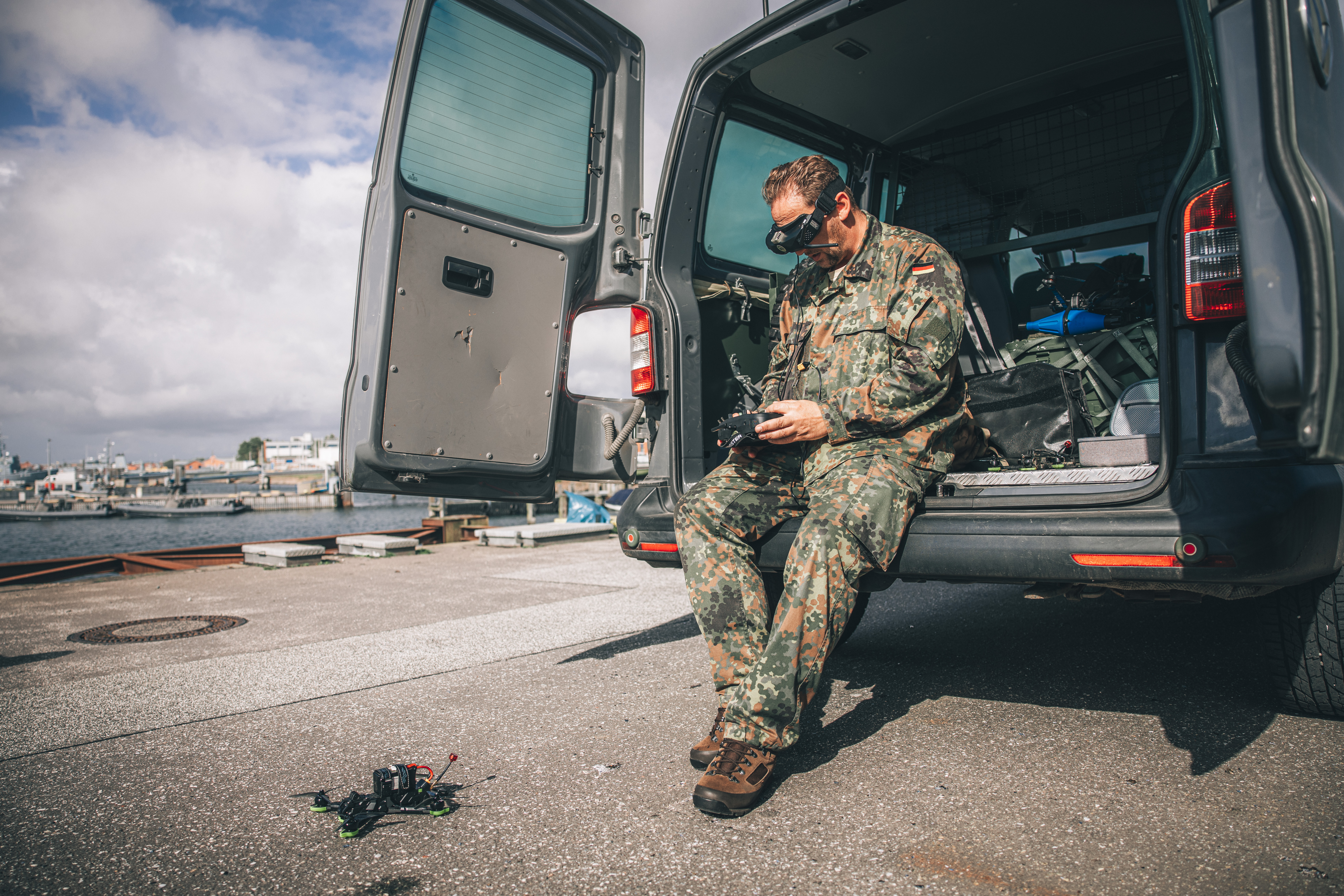 Ein Bundeswehrsoldat lehnt an die geöffnete Heckklappe eines Transporters. Er trägt eine FPV-Brille und hat ein Bediengerät in der Hand. Vor ihm liegt eine Drohne. Im Hintergrund ist eine Hafenanlage zu sehen.