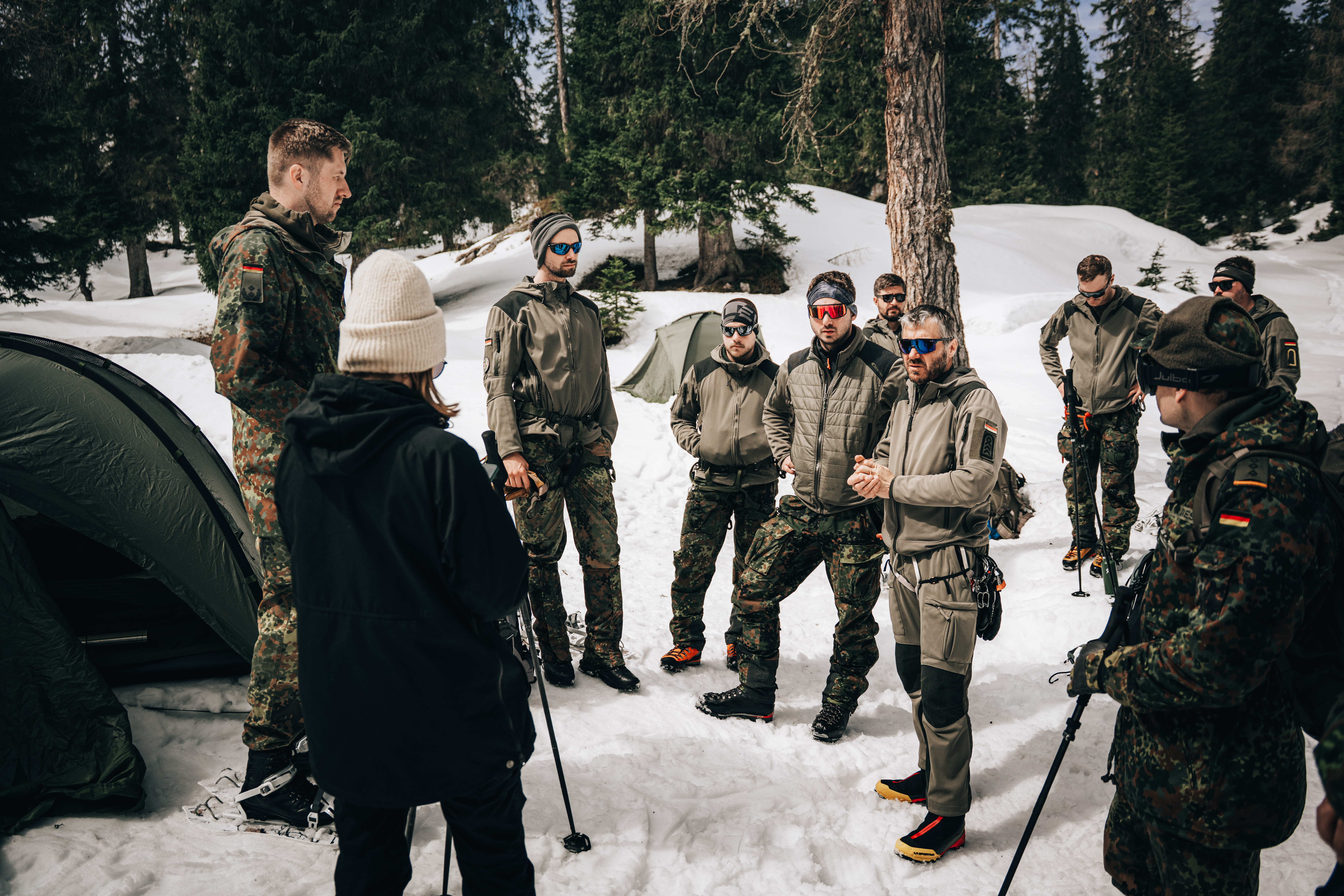 Eine Gruppe von Soldaten und Zivilisten steht im Kreis vor einer schneebedeckten Waldlandschaft
