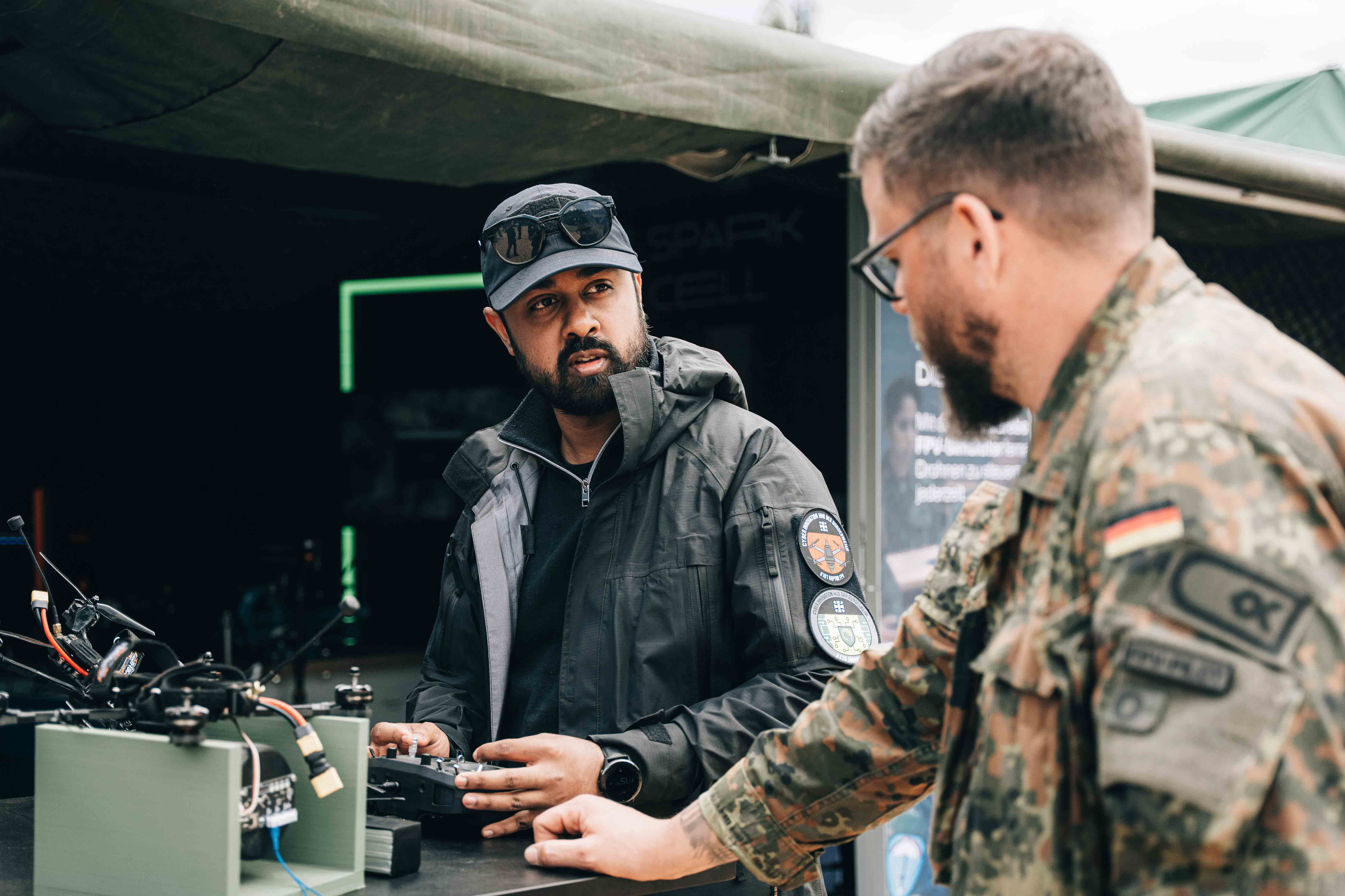 Ein Soldat und ein Zivilist unterhalten sich an einem Stehtisch, auf dem eine Drohne liegt, vor einem großen Bundeswehrzelt