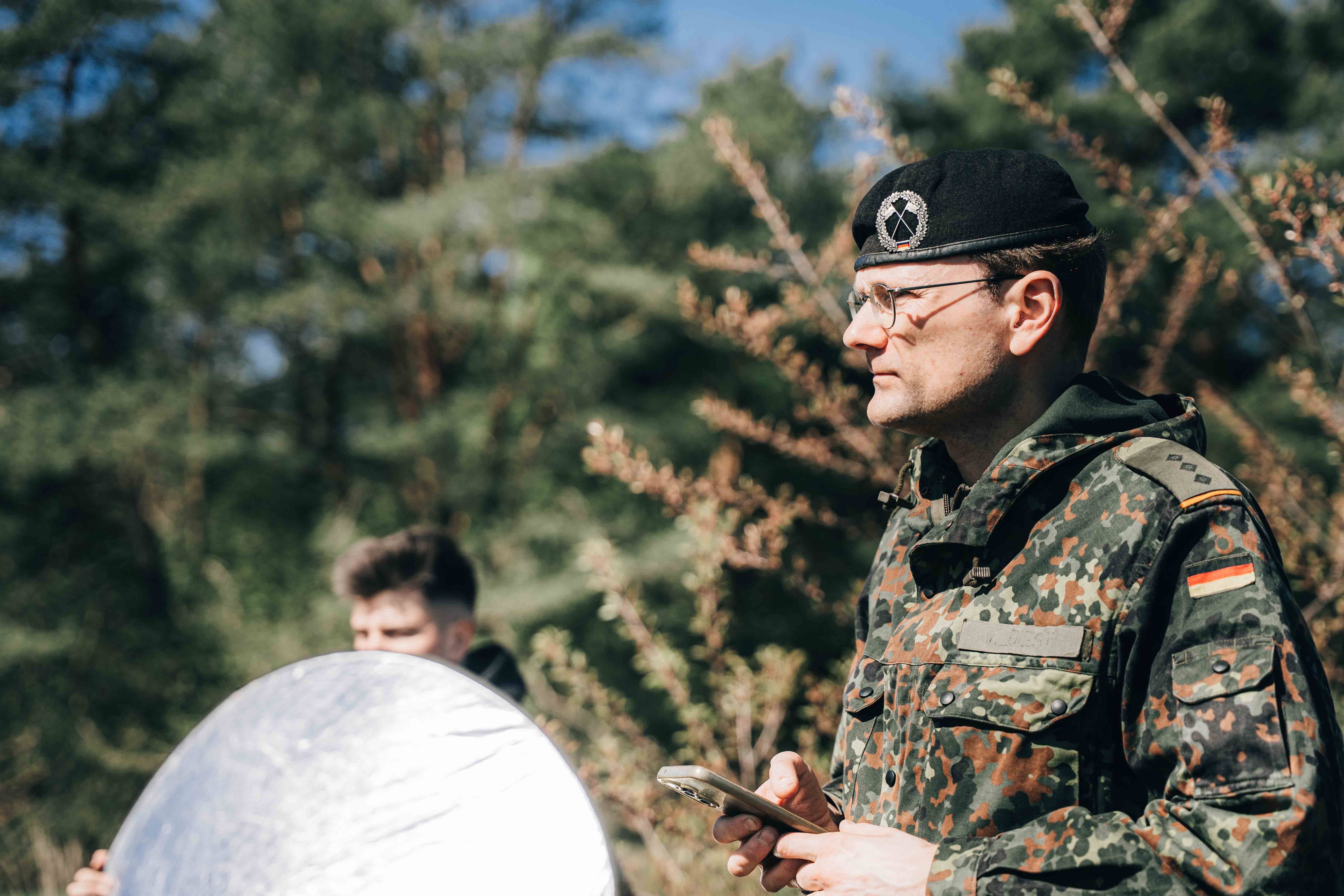 Ein Hauptmann der Heeresaufklärungstruppe mit Handy in der Hand steht rechts im Bild. Vor ihm ein Beleuchtungsspiegel. Im Hintergrund sind Baume und hohe Gräser.