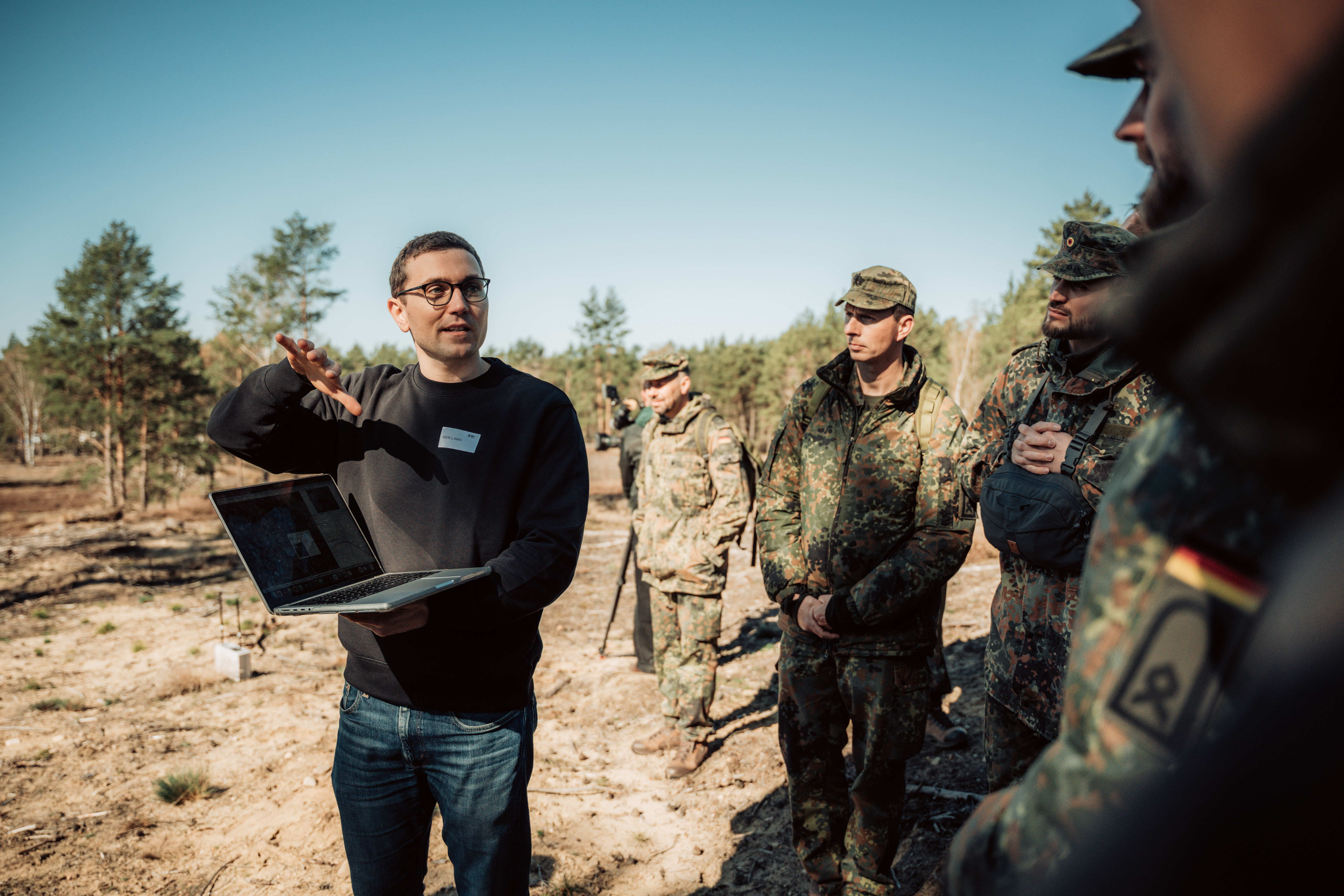 Ein Zivilist steht auf einer sandigen Freifläche in Brandenburg. Er hält einen Laptop in der Hand und erklärt umstehenden Soldaten seine Idee.
